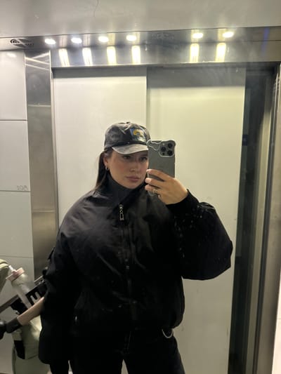 A person is taking a selfie in an elevator. The person is wearing a black jacket, a black turtleneck, and a gray baseball cap. The person is holding a smartphone in their left hand. The elevator has a mirrored wall and metal accents. The location is Селекционер-2 Снт, Russia.