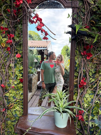 A vertical, reflected medium shot shows a couple kissing in front of a mirror adorned with artificial ivy and red flowers. The mirror is framed by a dark, ornate wooden border.
The couple, a man and a woman, are reflected in the mirror. The woman, with blonde hair, is wearing a floral dress and holding a pink smartphone, appearing to take a selfie. The man, wearing a green t-shirt with a red heart graphic and a black watch, is looking at her. Their faces are close, suggesting an intimate moment. The red heart graphic on his shirt contains white text that is not fully legible.
Behind the couple, the reflection shows an outdoor setting with trees, a wooden structure, and a blue sky with some clouds. The overall impression is of a warm, pleasant day. The scene is set outdoors, possibly in a garden or patio area, given the presence of plants and wooden furniture.
In the foreground, to the right of the mirror, a spider plant in a pale mint green pot is visible. The artificial ivy and red flowers surrounding the mirror suggest a decorative arrangement, perhaps at a cafe or an outdoor event. A black crow-shaped ornament is also visible at the top left of the mirror frame, adding a whimsical touch.
The image is taken at an angle, with the reflection appearing upside down relative to the viewer's perspective. The time of day appears to be daytime, indicated by the bright sky and shadows. The location context is Nizhnyaya Shilovka, Russia.