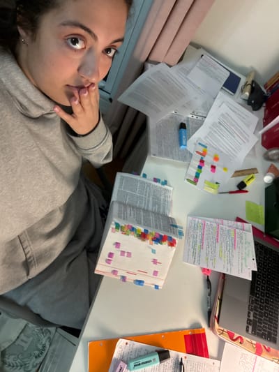 The image depicts a young adult female seated at a cluttered desk within what appears to be a residential room in Neuilly-sur-Seine, France.

The female subject occupies the left side of the frame, looking directly towards the camera with a wide-eyed expression. She has dark hair, a light-to-medium complexion, and a small silver stud earring in her left ear. She wears a grey hooded sweatshirt (hoodie) and matching grey sweatpants. Her right hand is raised, with fingers lightly touching her chin and lower lip; her fingernails have a light pink polish.

The desk surface, primarily white, is extensively covered with numerous study materials. These include multiple stacks of printed papers or open books, many heavily marked with colorful sticky notes/flags in shades of pink, blue, green, yellow, and orange along their edges. Several loose sheets of paper, some with handwritten notes and highlighting in various colors, are scattered across the desk.

Specific objects on the desk include:
*   Multiple highlighters (one light blue, one yellow, both appearing to be Stabilo Boss brand).
*   A red pen or marker.
*   A small wooden or brown ruler.
*   An open notebook or binder with extensive handwritten notes, highlighted in pink and yellow.
*   A silver-colored laptop, possibly a MacBook, resting on a base adorned with a pink flamingo pattern, visible on the right side of the desk.
*   A dark green binder or folder.
*   Several red-spined books or binders stacked vertically on the far right.
*   A dark blue folder or binder.
*   A small, clear cylindrical container and other small objects, possibly cosmetics or stationery.
*   An orange folder is visible at the bottom of the frame, alongside more papers and another light blue highlighter and a black pen.

In the background, behind the subject and the desk, light-colored walls are visible. Pink or peach-colored curtains partially obscure a window area along the top left of the frame. The overall lighting suggests an indoor setting, possibly with a mix of natural and artificial light.