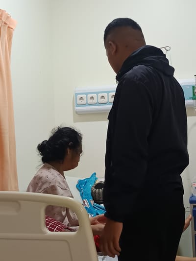 In a hospital room in Karangtengah, Indonesia, a man in a black jacket stands facing away from the camera, his back to the viewer. He appears to be looking at an elderly woman seated on a hospital bed. The woman is wearing a light pink patterned shirt and her hair is pulled back in a bun. She is facing the man, her head slightly bowed. A blue blanket with a cartoon pattern is visible on the bed. The room features medical equipment, including electrical outlets on the wall and a bed rail in the foreground. A peach-colored curtain is partially visible on the left side of the frame.