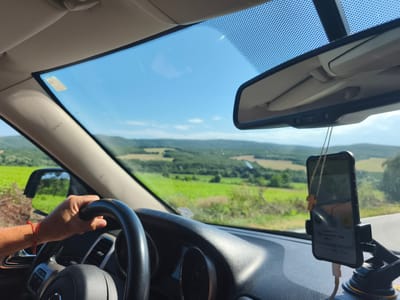 The image depicts the interior of a car, seen from the perspective of the driver. A hand, adorned with a red bracelet, is visible gripping the steering wheel. The dashboard features a smartphone mounted on a suction cup holder, displaying a navigation app. A small wooden cross hangs from the phone. Through the windshield, a scenic rural landscape unfolds, with green fields and rolling hills under a clear blue sky. The ambient light suggests it is daytime. There are no other individuals or specific city landmarks of Banya, Bulgaria, identifiable in this image; it shows a general view of a road trip through a countryside.