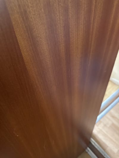 The image shows a close-up of a dark brown wooden surface, possibly a tabletop. The wood grain is visible, running diagonally across the frame. In the upper portion of the image, blurred metallic or light-colored elements suggest part of a structure, possibly a table leg or chair. The background is further blurred, showing a light-colored surface, possibly a floor. The location in Hereford, United Kingdom, is not discernible from the visual data. There are no people or subjects. The object is a wooden surface. There are no observable actions or interactions. The environment is undefined beyond the immediate surroundings of the wooden surface.