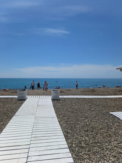 The image depicts a sunny day at a pebble beach in Krinitsa, Russia. The vast, clear blue sky occupies the upper two-thirds, with minimal wispy white clouds. A calm, deep blue-green sea extends across the middle ground to a flat horizon line. The foreground and midground consist of a coarse pebble beach, dark gray and brown in color. A central white wooden walkway, constructed from parallel slats, leads from the bottom center towards the sea. Two white wooden planter-like structures flank the walkway in the immediate foreground. Another section of white wooden path is visible entering from the bottom right.
Several individuals are present on the beach and in the water. A group of four adults is walking towards the sea along the central walkway's end, with one male carrying a black bag, a female in a dark dress, an older female in a white dress and straw hat, and a person in dark swim trunks. To the left, two individuals are seated on the pebbles near the water's edge. Further right, a person in a white t-shirt stands facing the sea, while others are swimming or lounging in the water and on the beach. One individual is paddling a small red inflatable craft near the shore on the right. The corner of a white structure is visible on the far right edge of the image near the horizon.