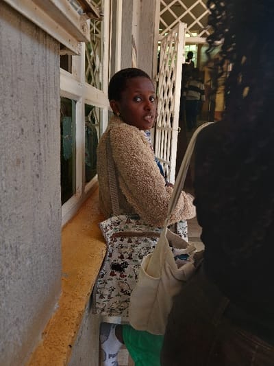 The image captures a scene within a hospital ward in Kenya. A young individual with dark skin, short dark hair, and small gold hoop earrings is the central subject, standing and looking directly at the camera. They wear a beige textured jacket, a white skirt patterned with various cartoon characters including black and white figures, green trousers, and white sneakers paired with white socks displaying a 'PUMA' logo on the heel. A beige corduroy shoulder bag is visible over their left shoulder. This individual stands beside a light grey concrete wall and a white-framed window with a yellowish-brown sill. To their right, an open white metal security gate with a lattice design leads to a background area where several indistinct figures are visible. In the immediate foreground, to the right, the back of a person with dark, curly hair and a dark garment is partially visible, facing away from the camera.