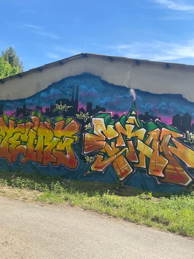 A concrete wall in Wysoka, Poland, displays graffiti art. The graffiti includes stylized lettering in shades of yellow, orange, and green, set against a painted backdrop of a city skyline with blue and pink hues mimicking a sunset or dawn. Adjacent to the wall, on the right, is a paved path with grass growing along the edge. A blue sky with faint clouds is visible above the wall. The photograph is taken from a ground-level perspective.