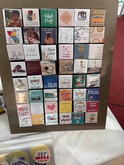 This image displays a brown rectangular board in Al-Ghizlaniya, Syria, affixed with forty small, square-shaped items, likely magnets or pins, arranged in a grid of approximately seven rows by six columns. The items exhibit a variety of designs, including photographs of individuals such as a smiling young boy, illustrations of abstract figures, hands, flowers, and religious motifs, alongside stylized Arabic calligraphy and English phrases. Notable English texts include "you won my heart," "PRAY," "KEEP CALM," "everything will be ok," and "You Complete Me!" The color scheme across the items is diverse. In the foreground, a white tablecloth is visible, partially obscured by clear plastic packaging containing yellow, cookie-like items, one marked with a red and white "GET WELL SOON" sticker. The background features a red carpet, suggesting an indoor setting.