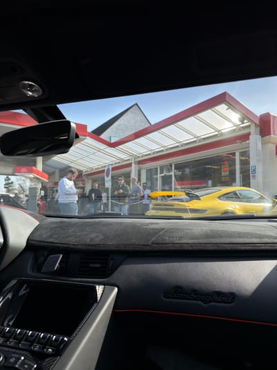 The image depicts a scene observed from the interior of a Lamborghini vehicle, specifically its black dashboard featuring an embossed "Lamborghini" script, red stitching, and carbon fiber trim around a dark display screen. A magnetic phone mount is attached to an air vent.
The external environment is a gas station in Meuspath, Germany, characterized by a red and white structure under a clear, paneled canopy. Fuel pumps are visible to the left. In front of the gas station store, approximately six to eight individuals are standing. One man in a white long-sleeve shirt is looking at a mobile phone. A bright yellow Porsche 911 GT3, distinguishable by its prominent rear wing, is parked facing away from the camera. A blue circular 'no stopping/parking' sign and a blue 'P' parking sign are visible. A price indicator displaying "1.84" is visible through the store window. Behind the gas station, a white, gabled building with a dark roof stands against a clear blue sky. The scene is illuminated by daylight.
