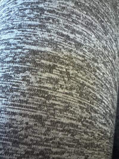 The image displays a close-up, textured view of a grey, marled knitted fabric. The fabric has a pattern of varying shades of grey and white, creating a heathered effect. The knitted loops are visible, forming horizontal lines with a slight curve that suggests the fabric is draped over a rounded surface. The lighting creates subtle shadows and highlights, emphasizing the texture of the knit. There is no discernible location in the city of Moelv, Norway, as the image is a close-up of a material and lacks any contextual environmental clues. No people, objects, actions, or interactions are visible.