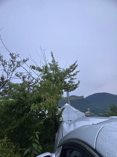 A silver car, partially visible in the foreground, is parked outdoors on a cloudy day. The car's hood is wet with raindrops, indicating recent precipitation. Behind the car, a large green cross stands prominently against a backdrop of lush green trees and a distant mountain range. The cross appears to be made of metal and is situated on top of a sloped, metallic structure. A few branches of a tree with green leaves are visible in the left side of the frame, partially obscuring the cross and the metallic structure. The sky is overcast and a light blue-grey color. The scene is likely located in a rural or mountainous area, possibly near a settlement given the faint buildings visible on the mountainside in the distance. The overall impression is one of a serene, somewhat somber, outdoor setting. There are no people visible in the image.