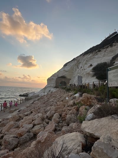 The image depicts a scenic view of Rosh HaNikra cliffs in the Mate Asher Regional Council, Israel, at sunset. The foreground is dominated by a rocky shoreline with large, irregularly shaped stones. A paved walkway runs alongside the shoreline, bordered by a metal railing. Several people are walking along this path, appearing to be tourists.
To the right of the walkway, a steep, white chalk cliff rises dramatically. At the base of the cliff, there's an entrance to a tunnel, with a few individuals gathered near it. Signs are posted along the walkway, some with informational text and pictograms.
The sea stretches out to the left, with gentle waves lapping against the shore. The sky is a gradient of soft blues and oranges, with scattered clouds catching the warm light of the setting sun, which is partially visible behind the cliffs. The overall atmosphere is tranquil and picturesque, highlighting the natural beauty of the location.