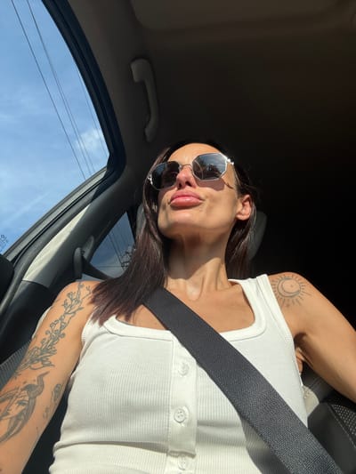 A woman with dark hair, wearing sunglasses, a white ribbed tank top, and a seatbelt, sits in the driver's seat of a car. She is looking up and to the right, her lips slightly pursed. Tattoos are visible on her left arm and right shoulder. The car interior is dark, with sunlight streaming in from the left. Outside the car window, the sky is blue with some clouds, and power lines are visible. The image was taken on a sunny day in Pordim, Bulgaria.