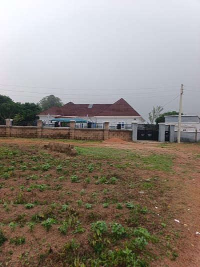 The image shows a modern house with a red tile roof, white walls, and dark windows, located in Reifild-Ist, Nigeria. The house is surrounded by a fence made of stone and metal. A black metal gate is visible in front of the house. The foreground of the image shows a field with sparse vegetation and red soil. The sky is overcast, suggesting cloudy weather. There are no people visible in the image. The scene appears to be a residential area.