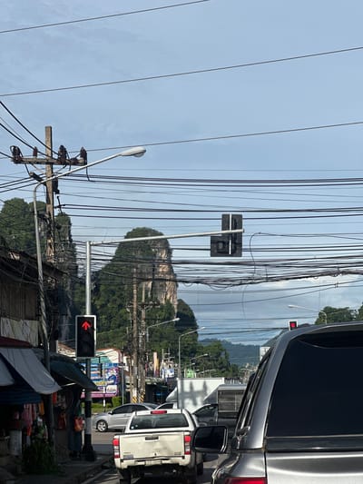 The image depicts a street scene in Plai Phraya, Thailand, characterized by a dense network of overhead electrical wires and a striking karst landscape in the background. In the foreground, a silver SUV is stopped at a traffic light, its rear visible. Ahead of it, a white pickup truck is also stationary. To the left, a traffic light displays a red upward arrow, indicating a permitted turn. A sidewalk with stalls selling merchandise lines the street, and a light pole with a street lamp stands nearby. Buildings with awnings are visible along the sidewalk. The background is dominated by lush green, rocky mountains with sheer cliffs. The sky is overcast, with visible cloud formations. Several traffic lights and street lamps are scattered throughout the scene, all partially obscured by the intricate web of power lines. The overall impression is one of a busy, yet visually distinctive, urban environment integrated with a dramatic natural backdrop.
