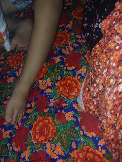 The image shows a close-up view of a person's leg and foot lying on a colorful floral-patterned fabric. The fabric features large red and orange flowers with green leaves on a blue background. To the right of the leg, other fabrics are visible, one with a dark floral pattern and another with a smaller, lighter floral pattern on an orange background. The scene appears to be indoors, possibly in a bedroom. There is no information to suggest the location is in the city of Aral, Kyrgyzstan.