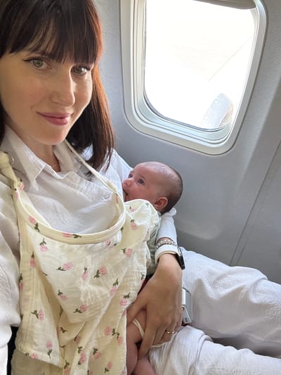 The image shows a woman lying down in what appears to be an airplane seat with a baby next to her, covered in a patterned blanket. The scene is likely inside an airplane cabin, evidenced by the presence of an airplane window showing part of the wing. The location is difficult to ascertain due to the lack of distinguishable landmarks. If the plane is flying in/out of Fiumicino, Italy the Fiumicino–Leonardo da Vinci International Airport (FCO) would be the relevant airport. The woman has dark hair with bangs, and the baby is lying on their side. The woman's hand is resting on the baby, appearing to be gently holding them. Both are wearing white clothing.