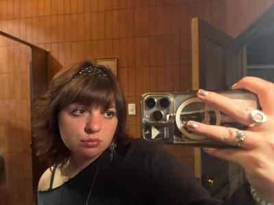 A young woman in a black top and dotted headband takes a selfie in a bathroom. She has short brown hair and a nose ring. Her phone is prominent, showing a clear case with a white circle inside, a "CHECKRA" sticker, and three camera lenses. Her hand holding the phone has silver nail polish and two rings. The background shows wood-paneled walls, a framed picture, a light switch, and a partially open door. The lighting is warm and dim.