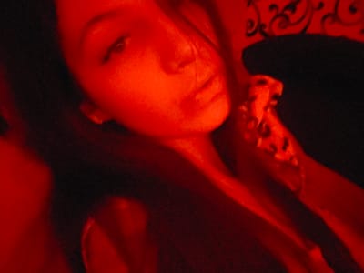 The image is a close-up, low-angle, self-portrait of a young woman against a dark background, bathed in red light. Her face is partially obscured by shadows, but her eyes, nose, and mouth are visible. She is wearing a leopard-print top. The scene appears to be indoors, possibly in a bedroom. The lighting creates a dramatic and intimate atmosphere. There is no indication of the specific location within Chystiakove, Ukraine.