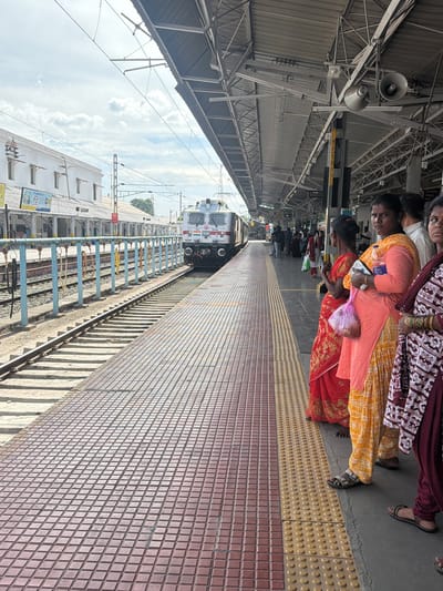 This image captures a scene at a train station in Thanjavur, India. The primary subjects are a group of people, predominantly women, standing on a platform. They are dressed in traditional Indian attire, including colorful sarees and salwar kameez. One woman is holding a pink plastic bag. The setting is a busy train station with a train visible on the tracks. The platform is lined with textured tiles and a tactile paving strip for accessibility. Overhead structures and electrical wires suggest an active railway environment. The background shows parts of the station building and the sky, which appears partly cloudy. The lighting suggests it is daytime. The people appear to be waiting, possibly for the train. No specific emotions are discernible, but the scene conveys a sense of everyday activity. Visible text includes markings on the train, such as "WAP-7 37210" and what appears to be the Indian flag. There is also some signage on the station building, but the text is not clearly legible.