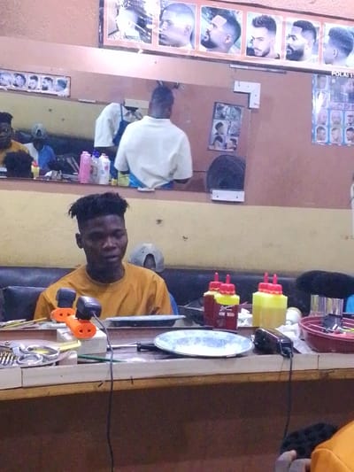 A young man with a high-top haircut wearing a mustard-colored shirt sits in a barber shop. In front of him is a counter with tools, including scissors, clippers, and bottles with red lids. Behind him, a mirror reflects the barber at work. A poster of men's haircuts is displayed above the mirror. The walls are painted a light brown color.