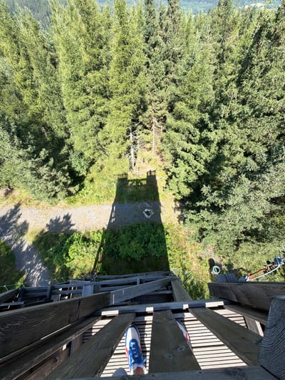 The image is a high-angle, downward view from a wooden observation tower in Świeradów-Zdrój, Poland, likely the Skywalk. The foreground shows the worn wooden planks of the tower's walkway and stairs, with the left foot of an individual wearing a blue and white athletic shoe and white socks visible on the walkway. Beyond the tower, a dense forest of coniferous trees covers the landscape. In the middle ground, a shadow of the tower falls on a gravel path and a clearing with a circular stone fire pit. To the right of the clearing, a green hose or rope is visible on the ground. Further in the distance, the forest continues, with the terrain appearing to slope downwards. The lighting suggests a sunny day.
