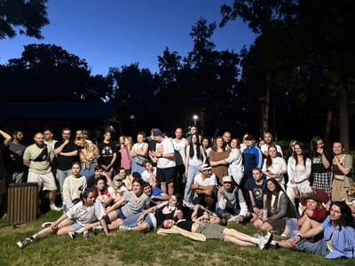 A large group of approximately 45 people is gathered outdoors in the Cukarica Urban Municipality, Serbia, possibly in a park or grassy area. The event seems to be taking place in the evening or late afternoon, as the sky is a deep twilight blue, and the lighting appears to be a mix of natural and artificial illumination.
The people are of varying ages and are dressed in casual attire. Many are standing, while others are sitting or lying on the grass. There's a relaxed and informal atmosphere, suggesting a social gathering or casual event. Some individuals are interacting with each other, and it appears that a few are posing for the photo.
In the background, there are trees and possibly a structure or covered area, which may be a pavilion or shelter. A trash can is visible on the left side of the frame. There is also a light pole. The overall impression is one of a communal gathering in a park-like setting.