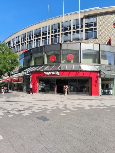 The image shows the entrance to a TK Maxx store in Wuppertal, Germany. The store is located on a paved pedestrian walkway with a patterned grey and white tile design. The building has a rounded facade with multiple floors. The upper floors are covered in a light-colored material with a diamond pattern and feature numerous windows with blinds. The TK Maxx store is at ground level, with a bright red facade and large glass windows displaying merchandise. Several people are visible on the walkway, including a man walking towards the store, a woman standing outside the entrance, and a few people inside the store. The sky is clear and blue, and a large green tree is visible to the left of the building. Red awnings with the TK Maxx logo are also visible along the front of the store.