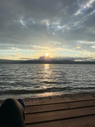 The image depicts a tranquil sunset scene over the body of water in Turgoyak, Russia. The sun is partially obscured by clouds, casting a warm, golden light across the rippling surface of the lake. The water is dark and appears calm, with gentle waves lapping at the pebble-strewn shore. In the foreground, the edge of a wooden dock is visible, with a person's legs and feet resting on it. The person is wearing dark pants and blue footwear. The distant shore is lined with dark, silhouetted mountains, adding depth to the landscape. The sky is filled with a mix of dark, brooding clouds and lighter, wispy formations, typical of a sunset. The overall impression is one of peacefulness and natural beauty.