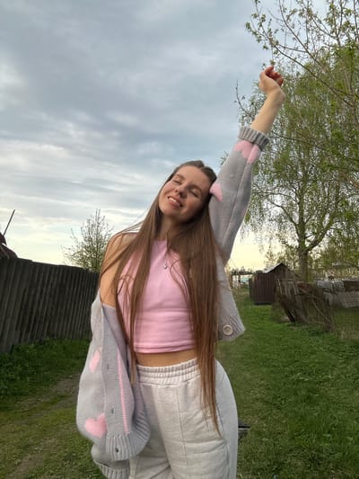 A young woman stands outdoors in what appears to be a rural or semi-rural setting, potentially within the area of Борщевка Малая, Russia, given the prompt's instruction. The woman is wearing a pink tank top, a gray sweater with pink heart designs, and light-colored sweatpants. Her right arm is raised towards a tree, while her left arm is bent at the elbow. She is smiling with her eyes closed. The environment includes a cloudy sky, a grassy area, trees, and some wooden structures, potentially sheds or outbuildings. There also appears to be a corrugated metal roof in the foreground. The image suggests a natural outdoor setting, possibly a garden or yard.