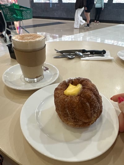 A close-up, eye-level shot shows a coffee and a pastry on a light brown table. The coffee, in a clear glass with a cardboard sleeve, is topped with cinnamon and cream. It sits on a white saucer. To the right, a golden-brown pastry, dusted with sugar, has a dollop of yellow cream in its center. The pastry rests on a white plate. A hand with bright red nail polish is visible on the right edge of the frame, reaching towards the pastry. In the background, out of focus, a shopping cart with green bags is visible. The floor is tiled with a reflective surface. The scene appears to be in a cafe within a shopping center, possibly located in the Toormina area of Australia.