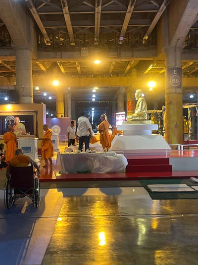 In a large, indoor, dimly lit space, possibly a covered market or transportation hub with exposed industrial-style ceilings and pillars, a ceremony appears to be underway. A golden statue of a seated figure, likely a religious icon, is centrally placed on a raised white platform with red steps leading up to it. Around the platform, several individuals in saffron robes, presumably monks, are gathered. One monk stands facing the statue, holding an object. Another monk is seated in a wheelchair in the foreground, facing away from the camera. Other individuals in casual clothing are also present, observing or participating. The floor is highly reflective, showing a golden sheen from overhead lighting. In the background, a large framed portrait of a person, also in saffron robes, is visible. A sign with the letter "E" and the number "88" is attached to one of the concrete pillars. The overall atmosphere suggests a formal or spiritual event.