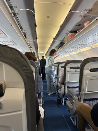 The image depicts the interior of an aircraft cabin. Passengers are seated in rows of airline seats. A woman stands in the aisle, possibly accessing the overhead compartment. Other passengers are seated. The environment is confined and typical of an airplane interior, featuring overhead compartments, seats, and blue carpeting. The location is likely an aircraft flying from or near Oberding, Germany, since the aircraft has signs in German.