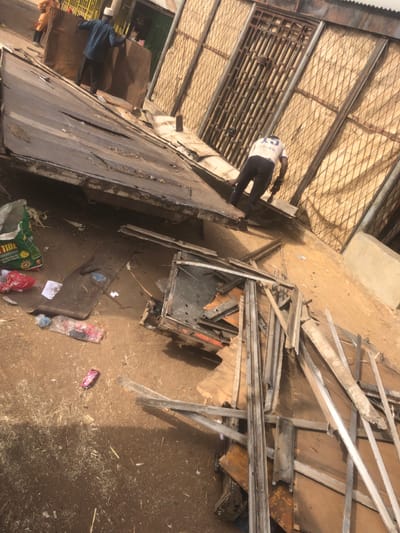 An outdoor scene in Tukur Tukur, Nigeria, depicts an area undergoing demolition or material salvage, characterized by scattered debris and active human presence. The ground is unpaved, consisting of light brown dirt or packed earth, with numerous wood shavings, discarded plastic items, and various scraps of metal and wood.
In the mid-ground, a large, dark, rectangular metal or wood panel, approximately 3-4 meters long, rests at an angle on the ground, indicating it has been detached from a structure. Its surface is weathered and appears dented in areas. To the immediate right of this panel, another large assortment of dismantled wooden and metal frames and beams lies on the ground, suggesting a collection of salvaged materials.
Three individuals are visible. In the mid-ground right, a person wearing a white shirt with the number "15" on the back and dark trousers stands with their back to the camera, bent over and seemingly interacting with the tilted panel. They appear to be holding a dark-colored tool, possibly a power tool or pry bar, against the panel. Further in the background to the left, a second individual, wearing a blue long-sleeved shirt and a light-colored cap, faces away from the camera while handling a large, flat, dark-colored sheet of material, possibly a piece of corrugated metal or wood. A third person, partially obscured, is visible further back, dressed in an orange-brown garment.
The background features a building facade with a prominent metal grate or lattice pattern, possibly a security feature or decorative element. A large, dark, heavily barred metal gate is integrated into this wall. To the far left, another building with a corrugated metal roof and what appears to be a small storefront is visible. A green and white "Tide" brand plastic bag is visible among the litter in the lower left foreground. The lighting suggests a bright, sunny day, with visible shadows on the ground.