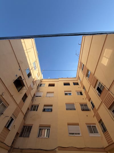 This media file shows a worm's-eye view of apartment buildings and the sky in Castelló De La Plana, Spain. The buildings are tall and painted a pale yellow color, with balconies and numerous windows. Some windows are open, revealing the interiors, while others are closed with blinds or shutters. There are also some visible exterior elements such as electrical wires and what appear to be satellite dishes or antennas. The perspective is dramatic, with the buildings seeming to stretch upwards towards a clear blue sky. The sun is shining, casting shadows on the building facades, suggesting it is daytime, likely morning or afternoon. There is no visible text in the image. The scene evokes a sense of urban living, looking up from a courtyard or narrow street. The overall impression is one of architectural geometry against the vastness of the sky.