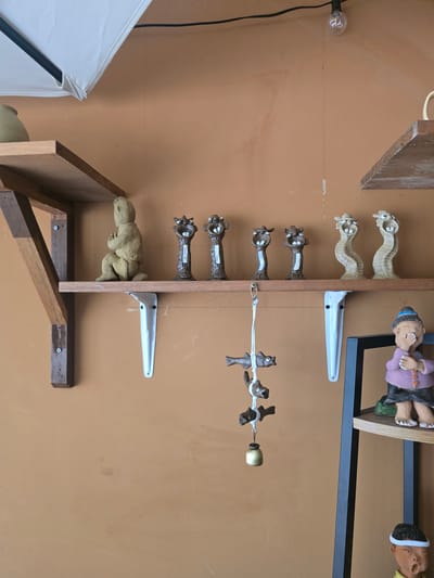The image depicts a display of various ceramic figurines on wooden shelves against an orange-brown wall. The top shelf holds a light beige, seated figurine resembling a person, and to its right, a row of eight small, abstract ceramic figures with open mouths and exaggerated features. Below this shelf, a string hangs down, adorned with three small wooden fish figures and a golden bell at the bottom.

To the right, on a lower shelf, a larger ceramic figurine of a person in a purple top and dark skirt is visible, with its hand covering its mouth. Further down, on another shelf, a head of a ceramic figurine with a yellow top and white headband is visible, with its mouth open.

The overall scene suggests a retail or display setting, possibly a souvenir shop or an artisan's studio. The image does not contain enough information to definitively place it in Gumi-si, South Korea.