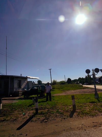 The image depicts a sunny day scene in the village of Kvarts, Russia. In the foreground, a dirt path with tire tracks leads to a grassy area where a small dog stands near a wooden post. To the left, a weathered wooden structure resembling a small house or shed is visible, with a tall pole and wires extending upwards. A dark-colored SUV is parked in front of the shed, and two men are standing near it. One man, wearing a light-colored shirt, is facing away from the camera, while another man, wearing a light-colored shirt and dark pants, is partially visible. The background shows a rural landscape with trees, a road, and more wooden posts with circular signs, suggesting a road crossing or intersection. The sun is high in the sky, casting long shadows.
