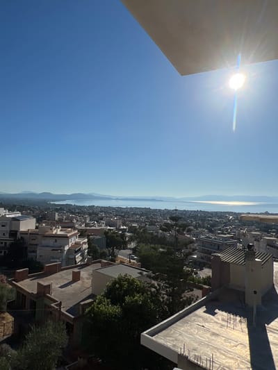 This is a daytime, high-angle photograph looking out over a coastal city. The foreground is dominated by the rooftops of several buildings, some appearing to be under construction with exposed rebar. The middle ground shows a dense urban landscape with various multi-story buildings, trees, and some visible streets. In the distance, a large body of water is visible, beyond which lie a range of hazy mountains under a clear blue sky. The sun is bright and positioned in the upper right quadrant of the image, causing a lens flare and casting long shadows across the rooftops. The overall impression is of a sunny day in a Mediterranean coastal town. The specific location is the Municipal Unit of Nea Makri, Greece, a coastal area on the eastern coast of Attica, known for its beaches and proximity to the Aegean Sea.