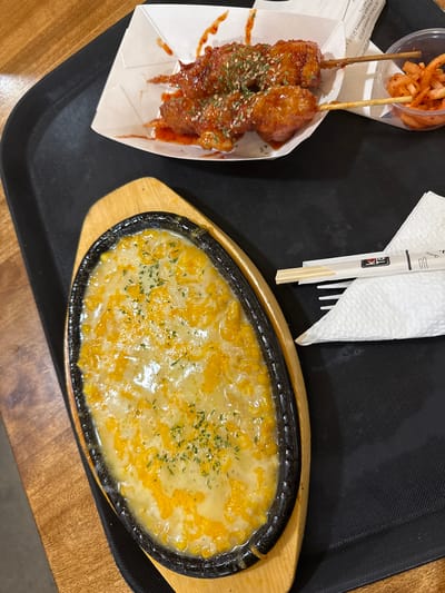The image shows a black tray on a wooden table, with several food items on it. On the left side of the tray, there is a paper container holding two chicken skewers covered in red sauce and garnished with herbs. Next to the skewers, there is a small container filled with what appears to be pickled radishes. To the right of these, there's a white napkin with a pair of chopsticks on top. On the right side of the tray, a sizzling plate with a cheesy dish is placed. The photograph was most likely taken indoors in Orlando, United States.