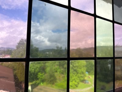 The image presents a close-up view of a multi-pane window, likely framed with dark lead or mullions, dividing the glass into numerous square and rectangular sections. The perspective is from indoors, looking out onto an outdoor scene in Lørenskog, Norway, during the daytime.
Through the upper panes, vibrant green foliage, possibly trees or bushes, is visible, with the glass itself exhibiting various textures and tints that distort and color the view. Some glass sections in the top row appear clear, while others have a crinkled or irregular surface. As the view shifts towards the middle and lower sections of the window, the blue sky with white clouds becomes prominent through several panes.
Many of the glass panes are distinctly tinted, displaying a range of colors including rose, pale pink, light green, lavender, grey, and varying shades of blue. These tints filter the natural light and the outdoor scene, creating a mosaic effect. Small droplets or imperfections are visible on the surface of many panes, contributing to a textured appearance and suggesting either condensation, light rain, or the inherent quality of the glass.
The scene is bright, indicating daylight. No people or text are visible, and there is no specific activity taking place beyond the observation of the outdoor environment through the unique window. The distinct framing and varied glass types suggest an older or traditionally styled window.