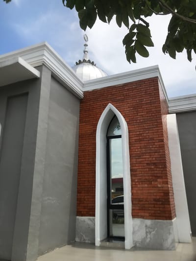 The image shows the exterior of a building in Meunasahceubrek, Indonesia, with a partially visible dome indicating it may be a mosque or related structure. A tree with green leaves is visible in the upper left corner. The building features gray concrete walls with layered moldings and a section of red brickwork. A narrow, arched window is set into the brickwork. The sky is visible in the background, with blue and white clouds. The photograph is taken from a low angle.