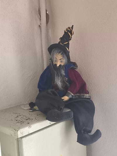 A doll representing a wizard, dressed in a dark pointed hat adorned with gold ribbon, a blue robe, and a burgundy vest with a gold trim, sits on the edge of a light-colored metal cabinet. The wizard doll has a long grey beard, a hooked nose, and appears to be made of cloth with painted facial features. Its legs are crossed, and it is positioned in an indoor environment against a textured, light-colored wall. To the left of the doll, on the cabinet, are a pair of glasses and some keys. A ribbed white pipe is visible on the wall behind the doll. The scene is located in Kuldīga, Latvia.