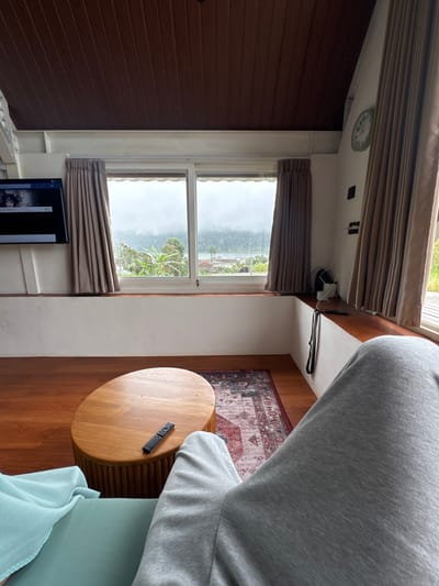The interior shot features a window with beige curtains framing a cloudy view. A dark wooden wall contrasts with the white window frame and wall space. A round wooden table sits on a hardwood floor near a patterned rug. A remote control rests on the table. Part of a couch with a blue blanket is visible in the foreground, along with a person wearing grey sweatpants. A television stands near the window. A clock can be seen on the ceiling.