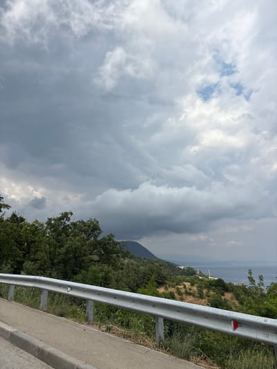 A guardrail is in the foreground of this image, separating a paved road from a vegetated hillside. Beyond the hillside is a body of water, which appears to be the sea, with a hazy horizon. The sky is filled with dramatic, dark clouds, suggesting an impending storm. The location is identified as Utios, Ukraine.