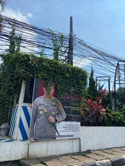 The image shows a poster of a uniformed Indonesian police officer standing in front of a green wall covered with vines. The poster is attached to a white wall with blue and white stripes. It is situated in a small, fenced-off area. The poster is framed in white, and has text in Indonesian language that reads "POLRI" and "NICO" on the bottom left corner. It also contains information about a police hotline. The poster is mounted on a metal pole. The environment is characterized by a large number of electrical wires. In the background, a red-leafed plant is visible next to the wall, with a large quantity of greenery in the distance. The overall scene is located in the city of Kecamatan Cimahi Selatan, Indonesia, at the coordinates: latitude: -6.905, longitude: 107.565.