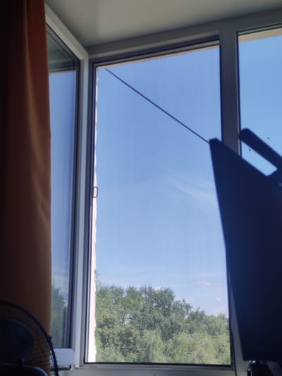 The image captures a view through a window, offering a glimpse of an outdoor environment and indoor elements. The window itself is a prominent feature, divided into sections by white framing. To the left, a portion of an orange curtain is visible, partially obscuring the view outside. Inside, a dark, angular object, possibly a screen or monitor, occupies the right side of the frame, casting a shadow. A thin, dark wire stretches diagonally across the upper portion of the windowpane.
The outdoor scene visible through the window consists of a bright blue sky with faint wisps of white clouds. Below the sky, a dense canopy of green trees stretches across the frame, indicating a leafy, possibly deciduous forest or park. The trees appear lush and full, suggesting a temperate season, likely spring or summer.
The lighting in the image suggests it is daytime, with sunlight illuminating the scene. The overall impression is one of a domestic interior looking out onto a natural landscape on a clear day. There are no individuals clearly depicted within the frame, nor are there any overt actions or interactions taking place. The scene is static and observational.
The provided location is Salavat, Russia. This information, while not visually evident within the image itself, contextualizes the scene as being situated in this specific geographical area. Without further visual cues, it is impossible to determine specific landmarks or characteristics unique to Salavat from the image alone.