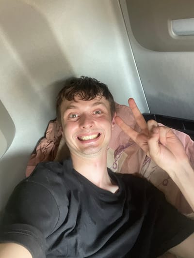 This media file is a selfie taken by a person lying down. The subject is a young man with short, dark, curly hair, a light complexion, and light-colored eyes. He is smiling broadly, showing his teeth. He is wearing a black t-shirt. His right arm is raised, and he is making a peace sign with his fingers. Behind him, there is a pinkish fabric with a subtle pattern, and a black garment that appears to be part of his clothing or bedding. The background suggests an indoor setting, possibly a train or a narrow room, with smooth, light-colored walls. The lighting is bright, suggesting daytime. The overall emotion conveyed is joyful and playful. There is no visible text in the image. The location context provided is Brachterbeek, Netherlands.