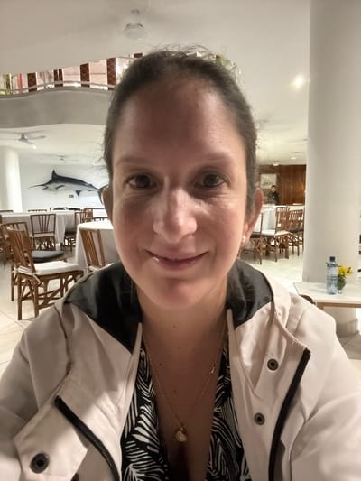 A person is taking a selfie in a restaurant in Ancón, Peru. The restaurant has white walls, wooden tables and chairs, and a painting of a swordfish on the wall. The person is wearing a white jacket over a black and white patterned shirt, and a gold necklace. In the background, there are other diners and staff. There is also a water bottle and flowers on a surface in the background.