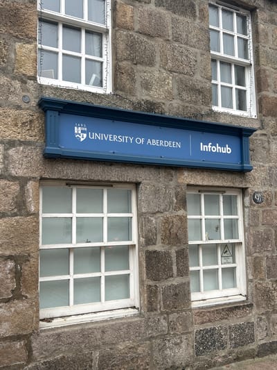 The image shows a stone building in Aberdeen City, United Kingdom. The building has three windows with white frames. A blue sign with the words "UNIVERSITY OF ABERDEEN" and "Infohub" is mounted on the building between the windows. One window has a "CCTV in operation" sticker. There is a small round plaque with the number "6" above one of the windows. A string of decorative lights is wrapped around the frame of one of the windows.