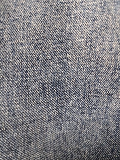 The image shows a close-up view of denim fabric. The texture is characterized by a diagonal weave pattern, with darker blue threads intertwined with lighter, possibly white or grey, threads. The lighting creates subtle shadows and highlights that emphasize the raised texture of the material. There is no indication of any specific location in the city of Tomsk, Russia, or any people, subjects, objects, or actions depicted. The image is purely a textured surface.