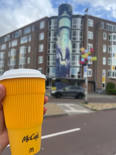 A hand holds a yellow McCafe branded paper cup against a blurred backdrop of a brown apartment building and a cloudy sky. The cup has a white lid and horizontal ridges. The McCafe logo is visible in black text on the cup, along with a small golden "M" logo. In the background, the apartment building features rows of windows and a mural painted on its side. The mural appears to depict a nighttime scene with a lighthouse. Colorful flags or decorations hang from a balcony on the building. A dark-colored vehicle is partially visible parked near the building. The scene suggests an outdoor setting, possibly a street or sidewalk, given the presence of pavement and a white line painted on it. The lighting and cloudy sky indicate it is daytime. The overall impression is a casual moment, perhaps someone enjoying a coffee while out and about in Leeuwarden, Netherlands.