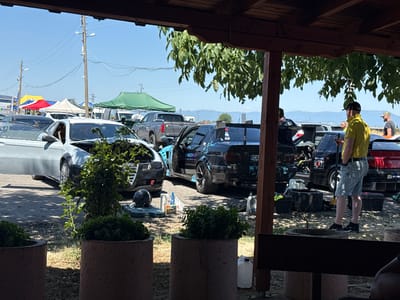 In Kaloyanovo, Bulgaria, under a clear blue sky and dappled sunlight filtered through tree leaves, a collection of modified vehicles is parked outdoors. The scene appears to be an informal gathering or preparation area for a motorsports event, possibly drifting, given the nature of the cars.
In the foreground, viewed from under a wooden overhang, are two large concrete planters containing green foliage. Beyond them, several cars are present. Closest to the viewer on the left is a light-colored sedan with its hood open, revealing its engine bay. To its right, a black sedan, heavily modified with a large rear spoiler and racing decals, also has its driver's side door open. This car has "RACING TO WIN" visible on its side. Further to the right, a red sedan is parked, its rear visible.
Several people are present. A man wearing a yellow polo shirt and denim shorts stands near the black sedan, facing away from the camera with his hands in his pockets. Another man in a black t-shirt and a camouflage baseball cap stands near the red sedan. Other individuals are partially visible or in the background.
Various items are scattered around the vehicles, suggesting maintenance or setup activities. These include tools, containers of liquid (likely oil or fuel), tires, and a helmet resting on the ground near the light-colored sedan. In the mid-ground, several tents and awnings are set up, some in bright colors like red, yellow, and blue, and others are green, providing shade. Electrical poles with wires run across the background, indicating proximity to an urban or developed area. Distant hills are visible on the horizon. The ground is a mix of asphalt and dirt.